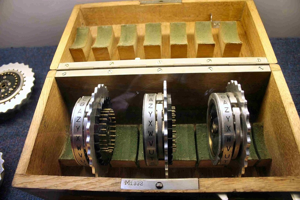 Three Enigma cipher rotors in their wooden storage box, with a fourth rotor lying beside it.